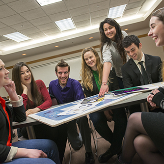 Business students sit around a table as one gestures to a poster laid out before them